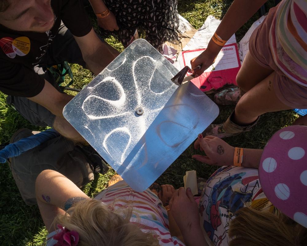 A square Chladni plate covered in white sand resonance patterns, surrounded by festival visitors.