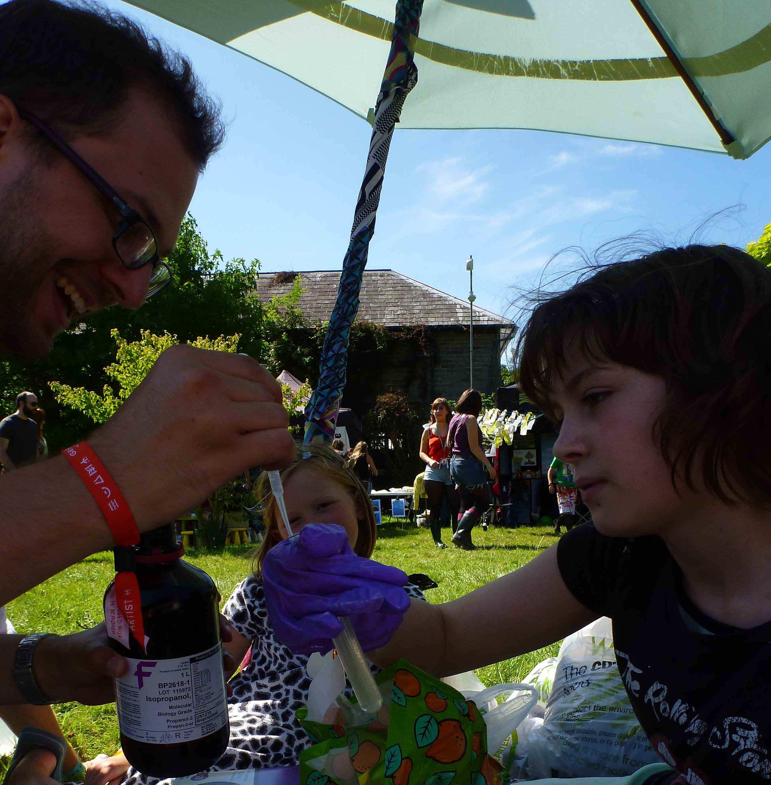 Outdoor festival demo under a parasol: a demonstrator pipettes isopropanol while two children in purple gloves hold a test tube, extracting DNA from a strawberry.