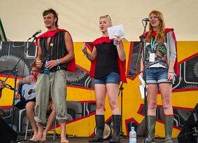 Three performers on a festival stage — one in a red cape, two in wellies reading lyrics — performing 'Physics Lab Blues' live.