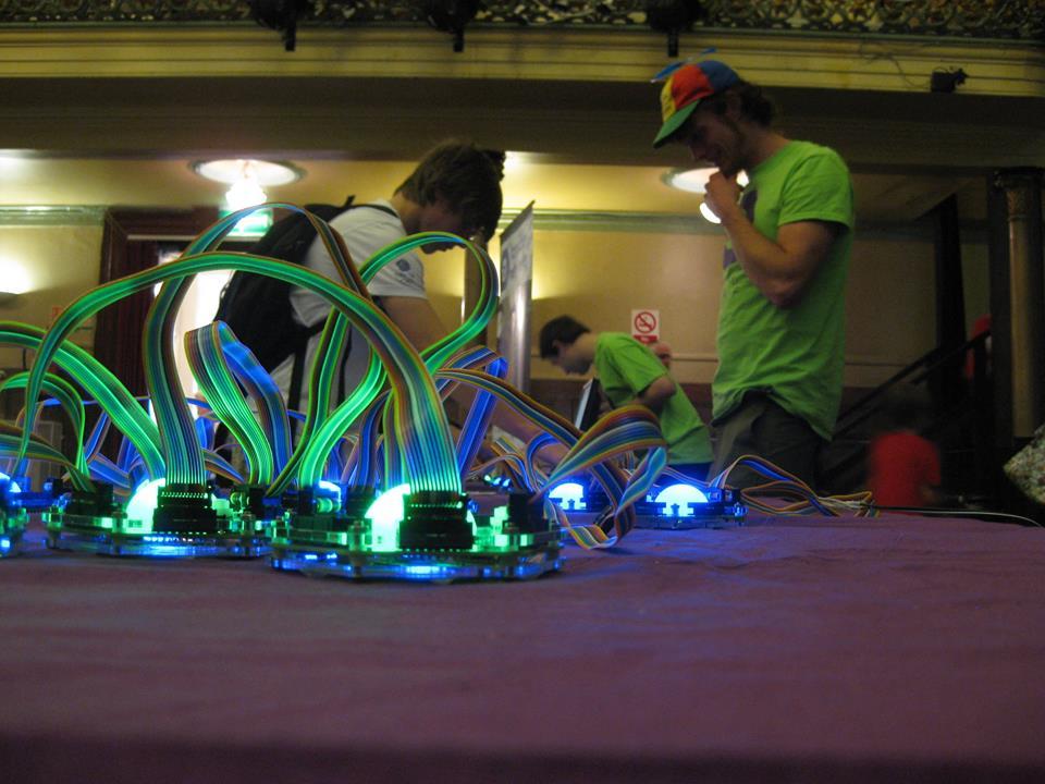 A table of Tangible Networks nodes glowing blue and green at a science festival, ribbon cables rising between them, with demonstrators in green project t-shirts behind.